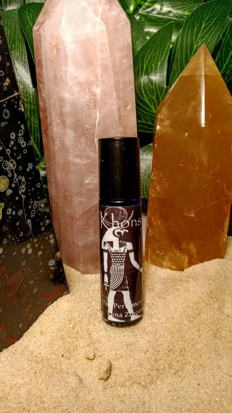 May include: A black roll-on bottle with a white label featuring an Egyptian goddess design and the text "Khonsu The Perfumers of Luna Zugar", surrounded by pink and orange crystals.