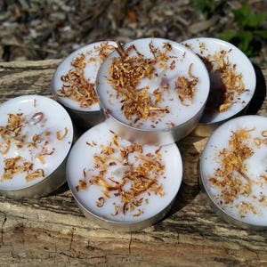 May include: Six white tea light candles with a decorative layer of brown flakes on top. The candles are arranged in a circle on a wooden surface.