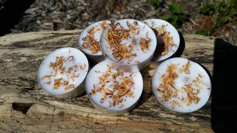 May include: Six white tea light candles with a decorative layer of brown flakes on top. The candles are arranged in a circle on a wooden surface.