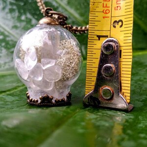 May include: A clear glass pendant necklace with a bronze-colored metal frame and a chain. The pendant contains small white crystals and a white, lacy material. The pendant is approximately 1 inch in diameter.