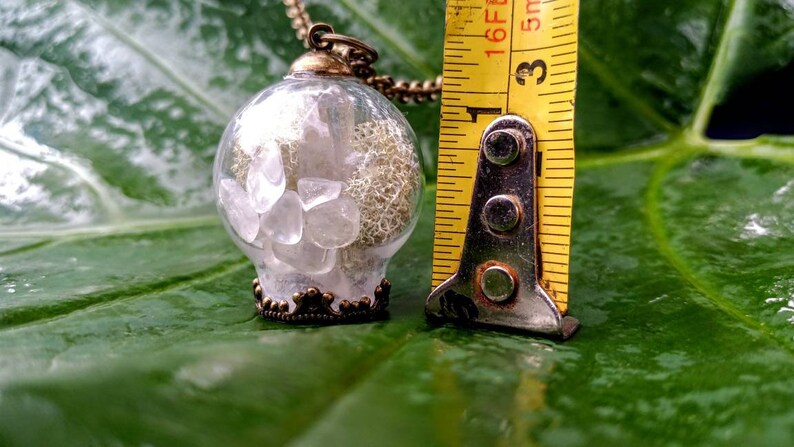 May include: A clear glass pendant necklace with a bronze-colored metal frame and a chain. The pendant contains small white crystals and a white, lacy material. The pendant is approximately 1 inch in diameter.