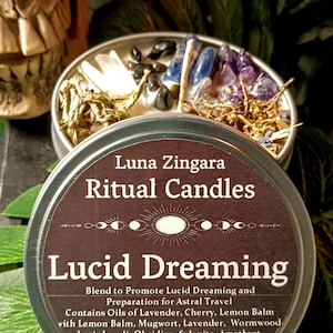 May include: A silver tin candle with a brown label that reads "Luna Zingara Ritual Candles Lucid Dreaming". The label also includes the text "Blend to Promote Lucid Dreaming and Preparation for Astral Travel. Contains Oils of Lavender, Cherry, Lemon Balm with Lemon Balm, Mugwort, Lavender, Wormwood, Lapis Lazuli, Obsidian, Selenite, Amethyst." The label also includes the website address "www.lunazingara.etsy.com". The candle is surrounded by crystals and herbs.