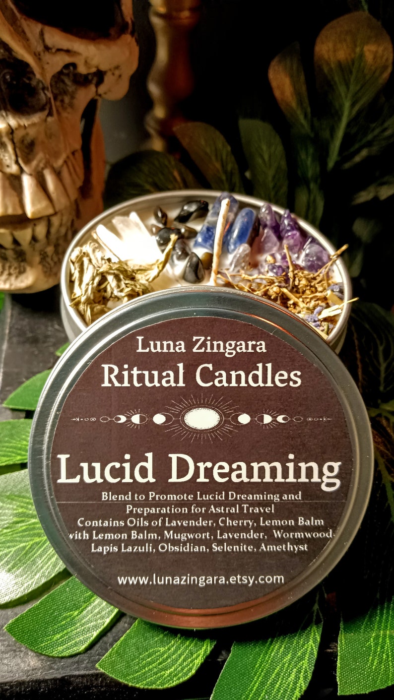 May include: A silver tin candle with a brown label that reads "Luna Zingara Ritual Candles Lucid Dreaming". The label also includes the text "Blend to Promote Lucid Dreaming and Preparation for Astral Travel. Contains Oils of Lavender, Cherry, Lemon Balm with Lemon Balm, Mugwort, Lavender, Wormwood, Lapis Lazuli, Obsidian, Selenite, Amethyst." The label also includes the website address "www.lunazingara.etsy.com". The candle is surrounded by crystals and herbs.
