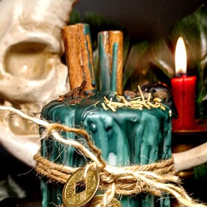 May include: A green and white candle with cinnamon sticks, twine, and two gold coins. A red candle burns in the background, with a skull and moon shape behind the candle.