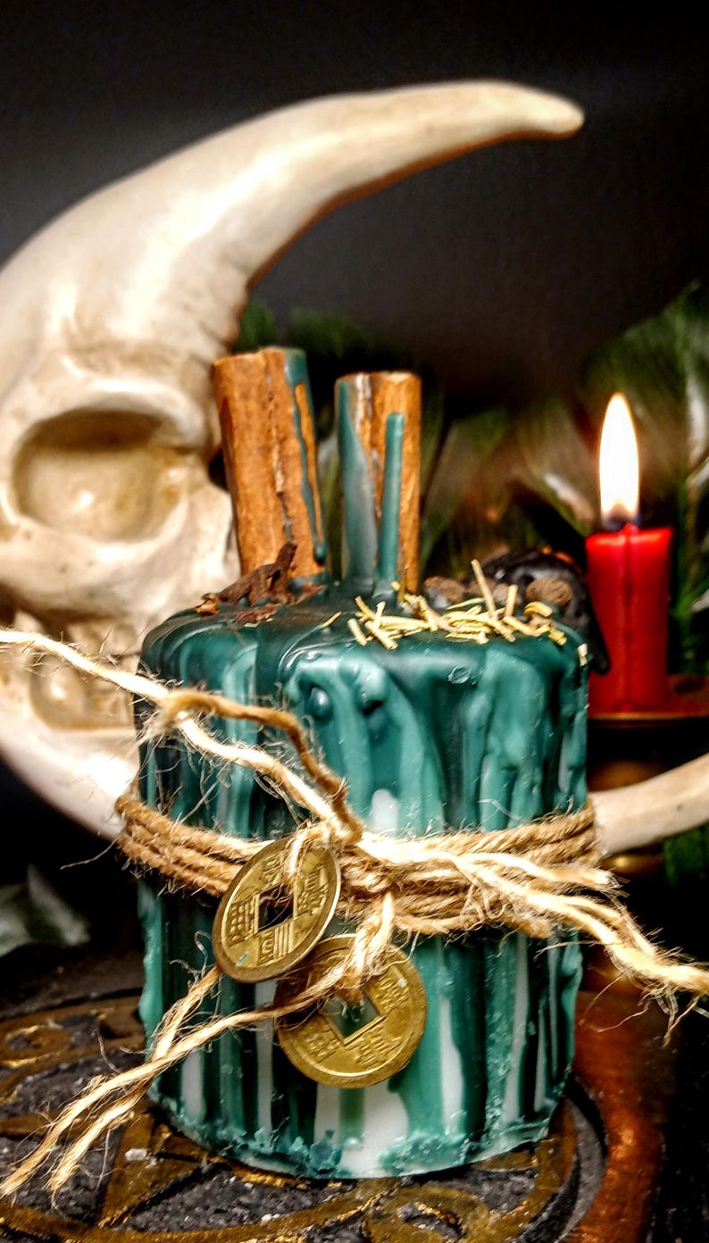 May include: A green and white candle with cinnamon sticks, twine, and two gold coins. A red candle burns in the background, with a skull and moon shape behind the candle.