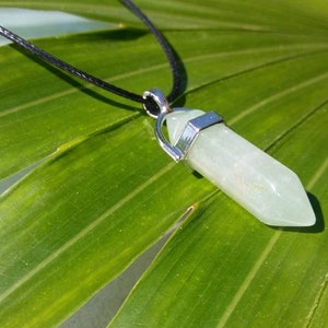 May include: A light green crystal pendant necklace with a black cord. The crystal is a hexagonal shape with silver-colored metal accents. The necklace is set against a green leaf background.