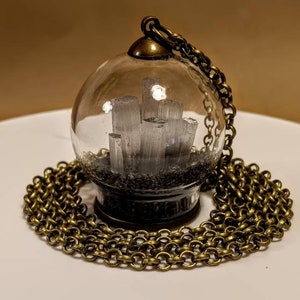 Peut inclure: Un collier avec un pendentif en forme de globe en verre et une cha&icirc;ne en laiton. Le globe contient un groupe de cristaux blancs sur une base noire.