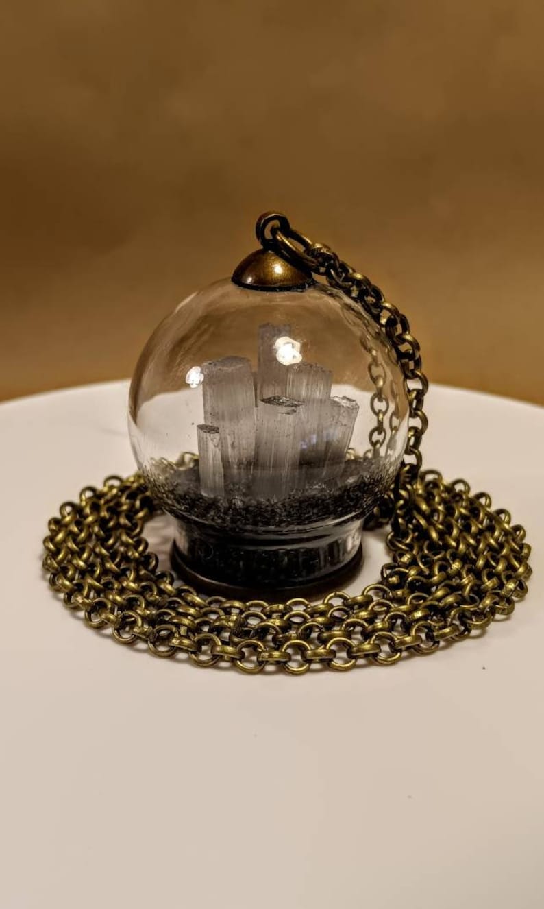 Peut inclure: Un collier avec un pendentif en forme de globe en verre et une cha&icirc;ne en laiton. Le globe contient un groupe de cristaux blancs sur une base noire.
