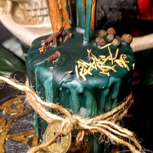May include: A dark green candle with cinnamon sticks and other herbs on top, tied with twine and a gold coin. The candle has a rustic, handcrafted appearance, suggesting a focus on natural elements and spiritual practices.