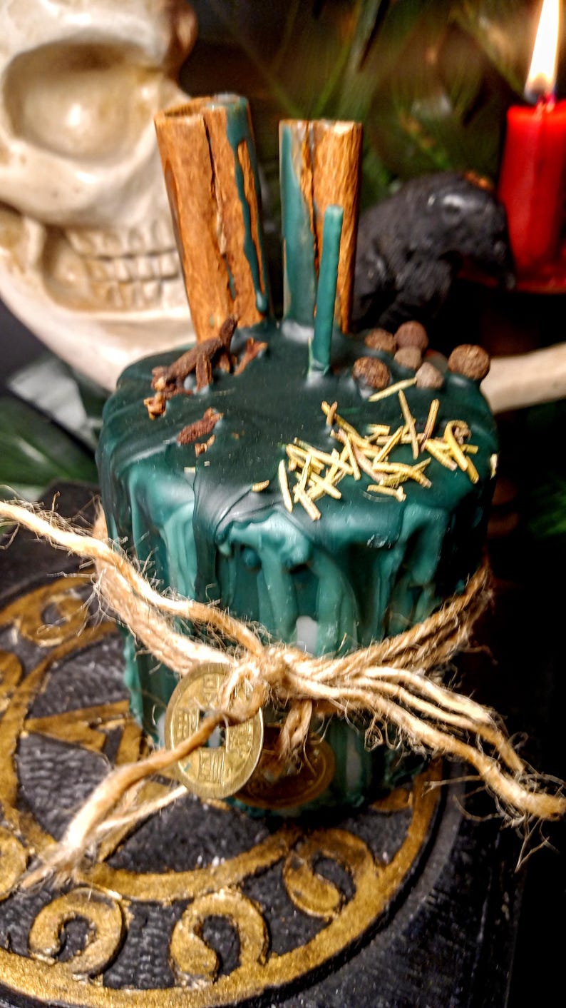 May include: A dark green candle with cinnamon sticks and other herbs on top, tied with twine and a gold coin. The candle has a rustic, handcrafted appearance, suggesting a focus on natural elements and spiritual practices.