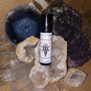May include: A small glass bottle of perfume oil with a white label that reads "Luna Zingara Perfume Oils Goddess Collection Persephone Garden". The bottle is surrounded by white and purple crystals.