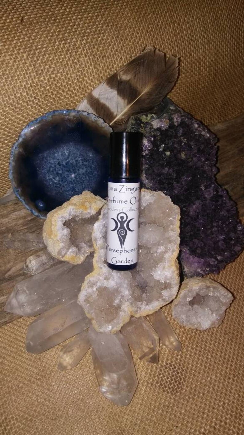 May include: A small glass bottle of perfume oil with a white label that reads "Luna Zingara Perfume Oils Goddess Collection Persephone Garden". The bottle is surrounded by white and purple crystals.