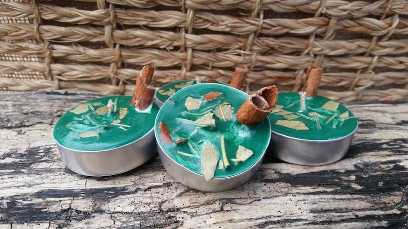 K&ouml;nnte beinhalten: Vier tealgr&uuml;ne Teelichtkerzen mit Zimtstangen und getrockneten Kr&auml;utern im Inneren. Die Kerzen befinden sich in Metallbeh&auml;ltern und stehen auf einer gewebten Oberfl&auml;che.