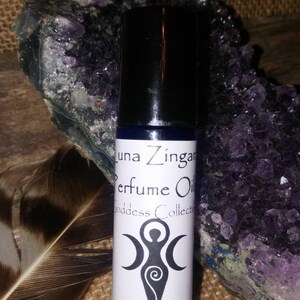 May include: A white bottle with a black cap and a white label. The label says "Luna Zingara Perfume Oil Goddess Collection Persephone's Garden". The bottle is sitting on a brown surface with a purple rock and a feather.