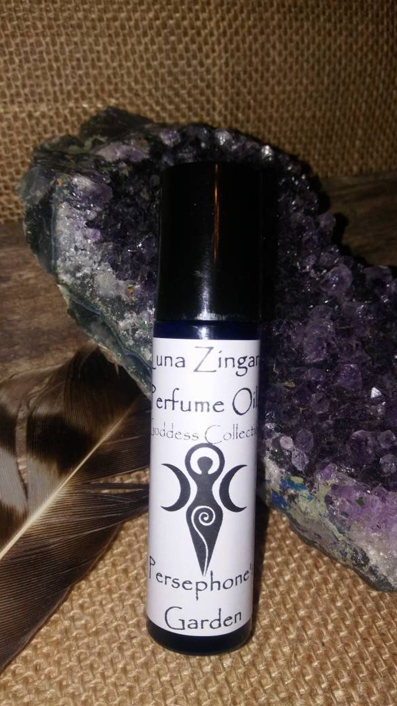 May include: A white bottle with a black cap and a white label. The label says "Luna Zingara Perfume Oil Goddess Collection Persephone's Garden". The bottle is sitting on a brown surface with a purple rock and a feather.