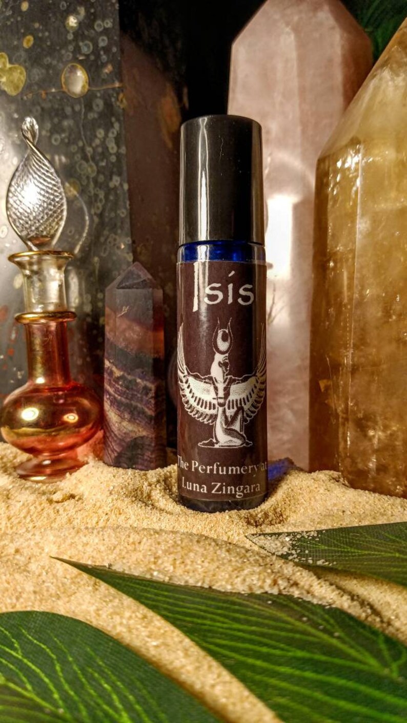 May include: A perfume bottle with a black cap and a brown label featuring the word "isis" and an illustration of a winged figure. The bottle is surrounded by crystals and sand.