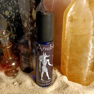 Puede incluir: Un frasco de vidrio azul con un tap&oacute;n negro y una etiqueta que dice "Anubis The Perfumer Luna Zimmera". La etiqueta presenta una ilustraci&oacute;n en blanco y negro de un dios egipcio con cabeza de chacal.
