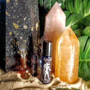 Puede incluir: Una botella de vidrio azul con una tapa plateada y una etiqueta que dice "Anubis The Protector Limited Edition" se muestra sobre una superficie arenosa con dos puntas de cristal, una rosa y una naranja, y una planta verde frondosa en el fondo.