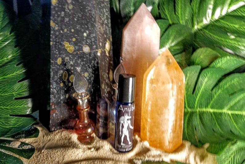 Puede incluir: Una botella de vidrio azul con una tapa plateada y una etiqueta que dice "Anubis The Protector Limited Edition" se muestra sobre una superficie arenosa con dos puntas de cristal, una rosa y una naranja, y una planta verde frondosa en el fondo.
