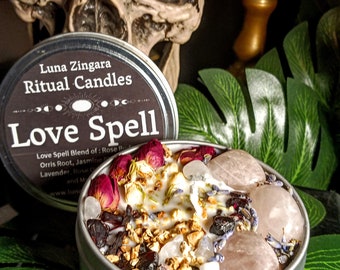 May include: A silver tin candle with a black label that reads "Luna Zingara Ritual Candles Love Spell". The candle is filled with white wax, rose petals, lavender, and crystals. The candle is sitting on a green leaf.