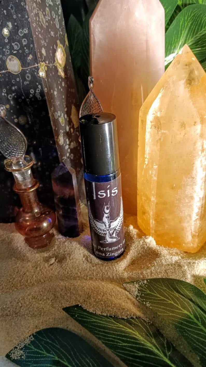 May include: A roll-on perfume bottle with the word "Isis" and a winged figure on the label. The bottle is surrounded by crystals and sand, with a backdrop of green leaves. The perfume is from Perfumery Ana Zingara.