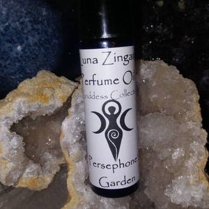 May include: A small, clear glass bottle with a black cap and a white label. The label says "Luna Zingara Perfume Oil Goddess Collection Persephone's Garden". The bottle is sitting on a white and brown crystal.