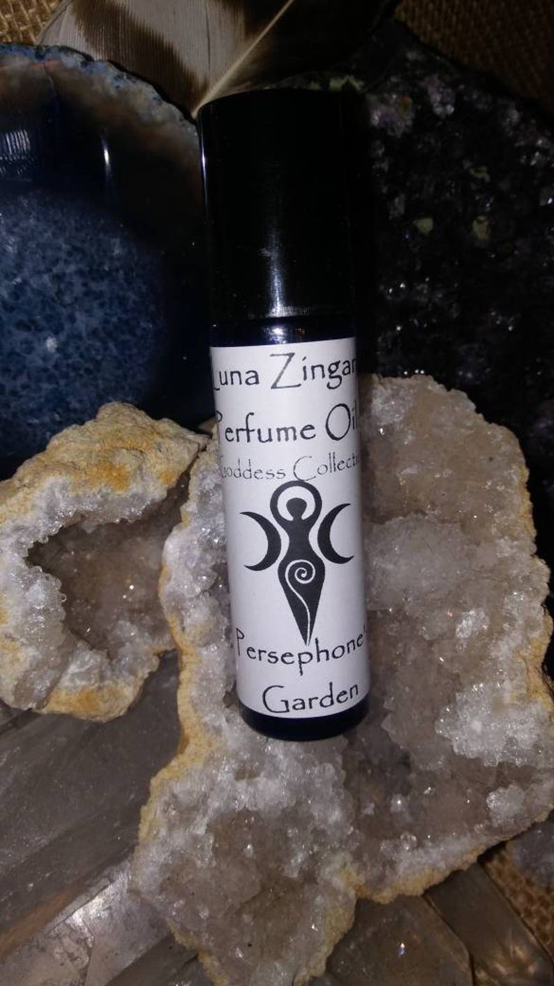 May include: A small, clear glass bottle with a black cap and a white label. The label says "Luna Zingara Perfume Oil Goddess Collection Persephone's Garden". The bottle is sitting on a white and brown crystal.