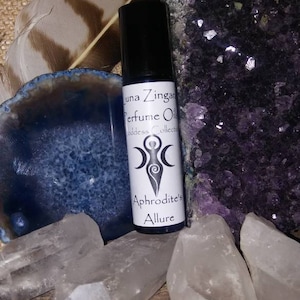 May include: A small, clear glass bottle with a black roll-on top. The bottle is labeled "Luna Zingara Perfume Oil Goddess Collection Aphrodite's Allure". The bottle is surrounded by crystals and a feather.