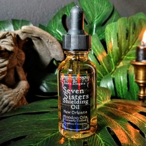 May include: A clear glass bottle of Seven Sisters Shielding Oil with a black dropper. The label reads "Conjure Oils by Luna Zingara, New Orleans Voodoo Oils, Curiously Crafted." The bottle sits on a green leaf with a lit candle in the background.