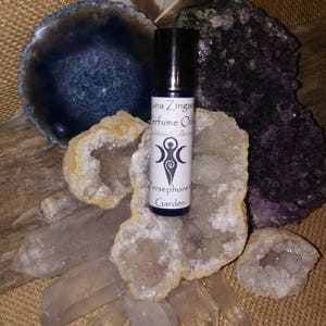 May include: A small glass bottle of perfume oil with a white label that reads "Luna Zingara Perfume Oil Goddess Collection Persephone Garden". The bottle is surrounded by crystals and a feather.