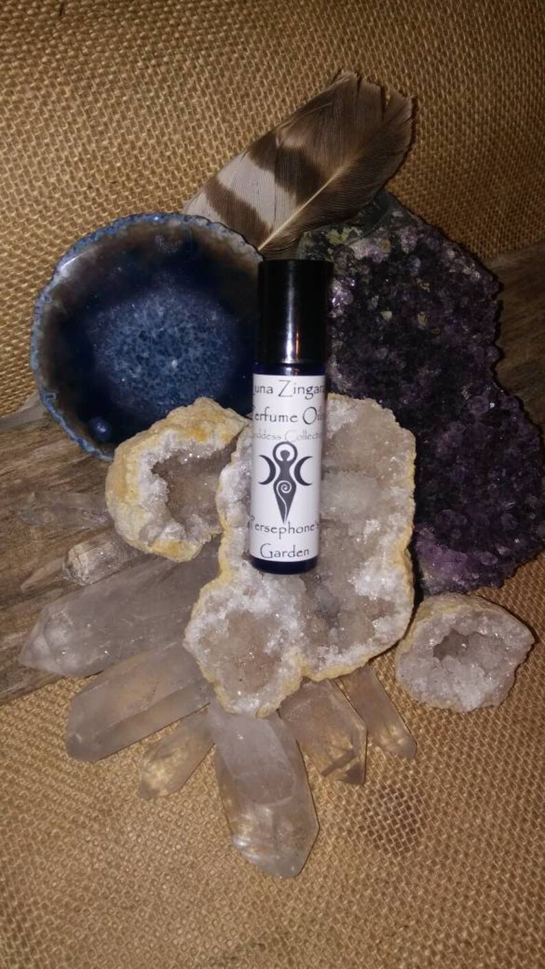 May include: A small glass bottle of perfume oil with a white label that reads "Luna Zingara Perfume Oil Goddess Collection Persephone Garden". The bottle is surrounded by crystals and a feather.