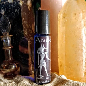Puede incluir: Un frasco de vidrio azul con tapa negra y una etiqueta que dice "Anubis The Perfumery Luna Zingaro". La etiqueta presenta una ilustraci&oacute;n en blanco y negro de un dios egipcio con cabeza de chacal.