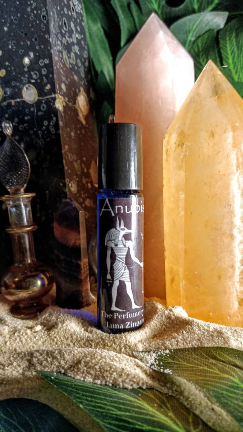 Puede incluir: Un frasco de vidrio azul con tapa negra y una etiqueta que dice "Anubis The Perfumery Luna Zingaro". La etiqueta presenta una ilustraci&oacute;n en blanco y negro de un dios egipcio con cabeza de chacal.