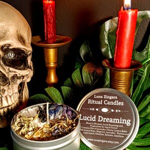May include: A silver tin candle with a label that reads "Luna Zingara Ritual Candles Lucid Dreaming Blend to Promote Lucid Dreaming and Preparation for Astral Travel Contains Oils of Lavender, Cherry, Lemon Balm with Lemon Balm, Mugwort, Lavender, Wormwood Lapis Lazuli, Obsidian, Selenite, Amethyst www.lunazingara.etsy.com". The candle is surrounded by green leaves and two lit red candles on gold candle holders.