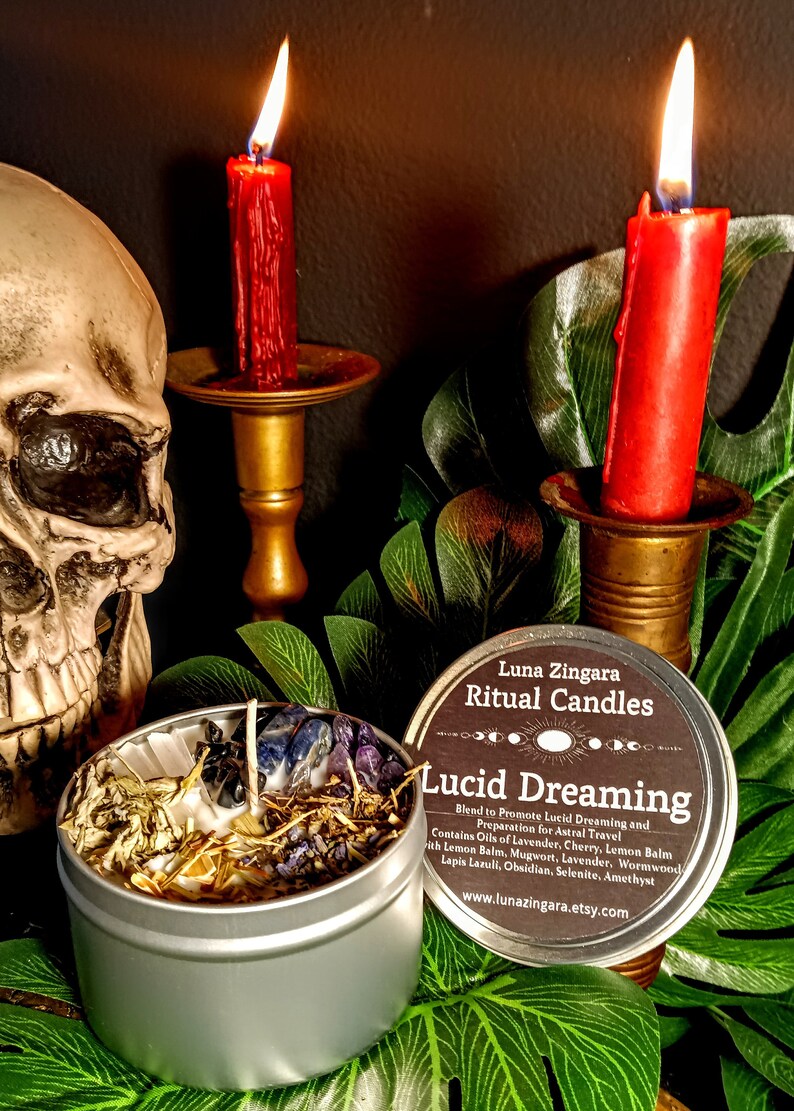 May include: A silver tin candle with a label that reads "Luna Zingara Ritual Candles Lucid Dreaming Blend to Promote Lucid Dreaming and Preparation for Astral Travel Contains Oils of Lavender, Cherry, Lemon Balm with Lemon Balm, Mugwort, Lavender, Wormwood Lapis Lazuli, Obsidian, Selenite, Amethyst www.lunazingara.etsy.com". The candle is surrounded by green leaves and two lit red candles on gold candle holders.