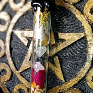 May include: A glass vial necklace with a black metal top. The vial is filled with dried herbs, flowers, and crystals. The necklace is hanging on a gold pentagram.