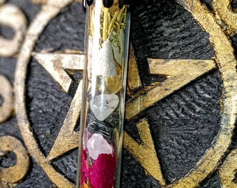 May include: A glass vial necklace with a black metal top. The vial is filled with dried herbs, flowers, and crystals. The necklace is hanging on a gold pentagram.