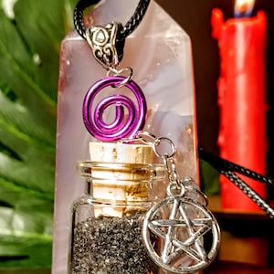 May include: A black glass bottle filled with black sand, with a cork stopper and a silver chain with a pentagram charm. The bottle is hanging from a black cord with a purple wire spiral charm.