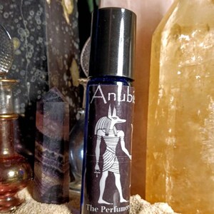 Puede incluir: Un frasco de vidrio azul con forma de rodillo con una tapa plateada y una etiqueta que dice "Anubis The Perfumery Luna Zingara" y presenta una ilustraci&oacute;n en blanco y negro del dios egipcio Anubis.