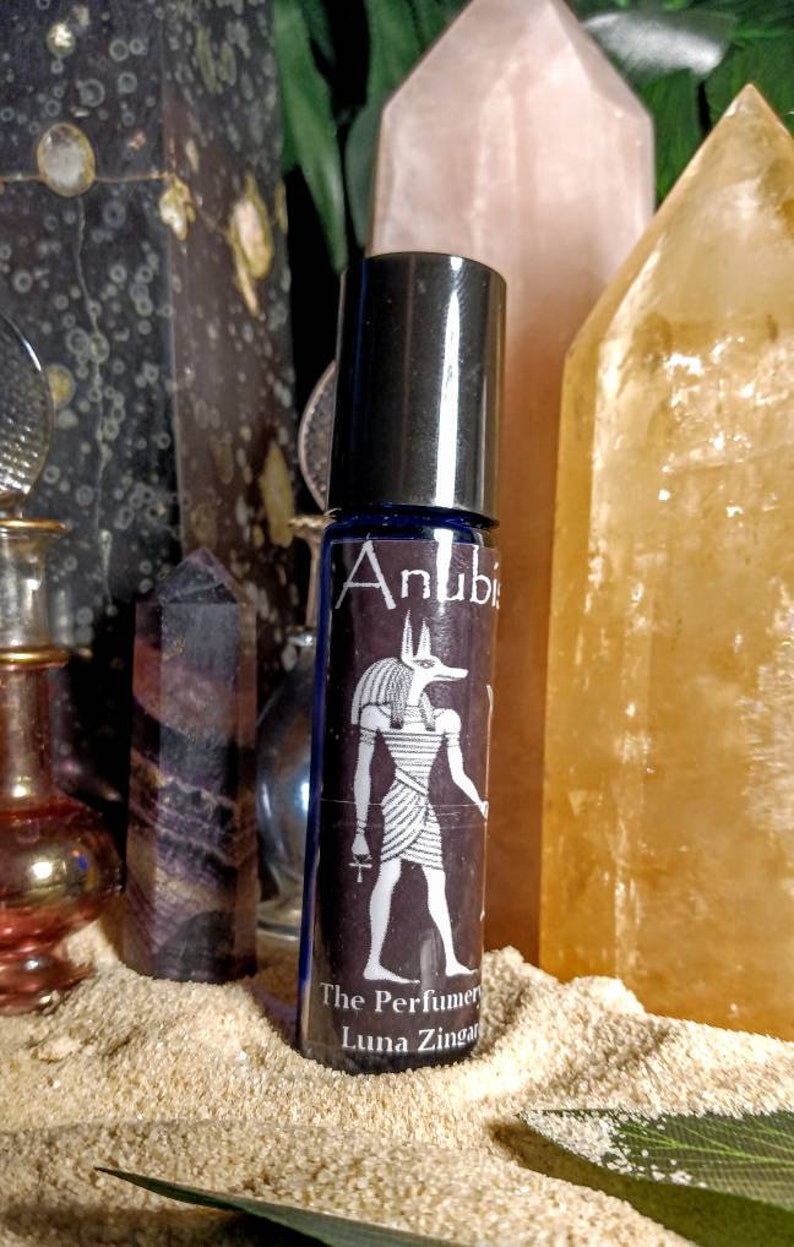 Puede incluir: Un frasco de vidrio azul con forma de rodillo con una tapa plateada y una etiqueta que dice "Anubis The Perfumery Luna Zingara" y presenta una ilustraci&oacute;n en blanco y negro del dios egipcio Anubis.