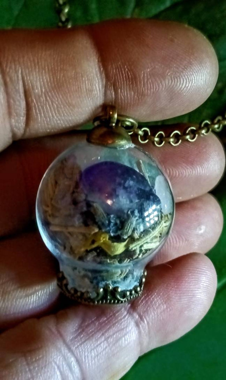 May include: A glass orb pendant necklace with a brass chain. The orb contains dried herbs, crystals, and a purple amethyst stone.