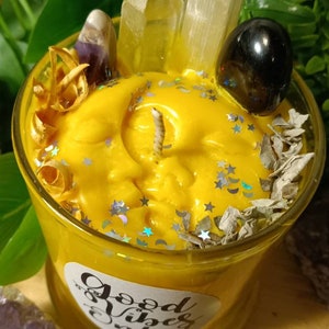 May include: A yellow candle with a moon and stars design, surrounded by crystals, dried herbs, and glitter. The candle is in a glass jar with a label that reads "Good Vibes Only".