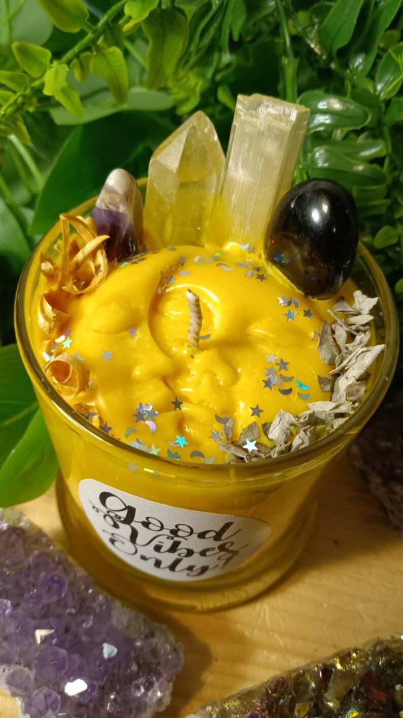 May include: A yellow candle with a moon and stars design, surrounded by crystals, dried herbs, and glitter. The candle is in a glass jar with a label that reads "Good Vibes Only".