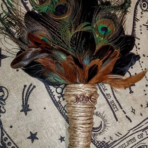 May include: A brown and black feather smudge stick with a triple moon symbol and a jute handle. The smudge stick is on a fabric background with a hand print design and stars.