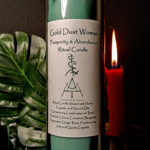 May include: A teal green glass candle with a gold label that reads "Gold Dust Woman Prosperity & Abundance Ritual Candle". The candle is decorated with twine and gold coins. The label also includes a description of the candle's ingredients, including herbs, crystals, and sacred oils.