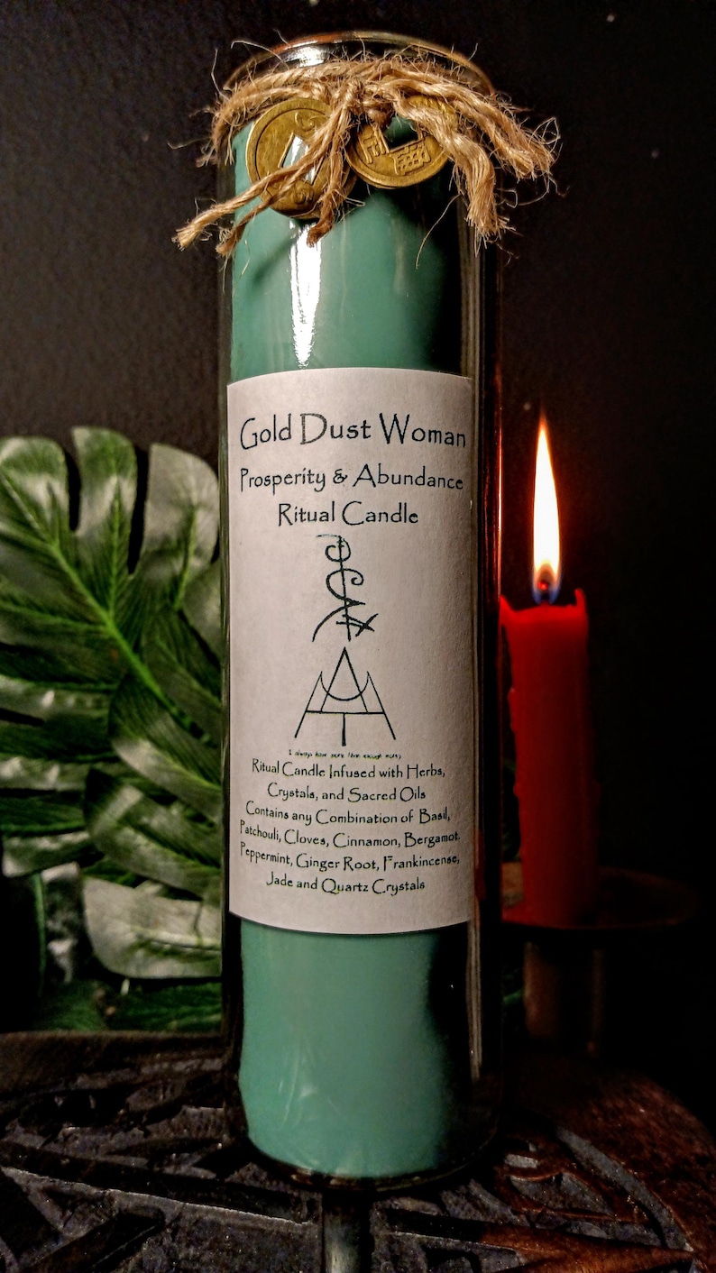 May include: A teal green glass candle with a gold label that reads "Gold Dust Woman Prosperity & Abundance Ritual Candle". The candle is decorated with twine and gold coins. The label also includes a description of the candle's ingredients, including herbs, crystals, and sacred oils.