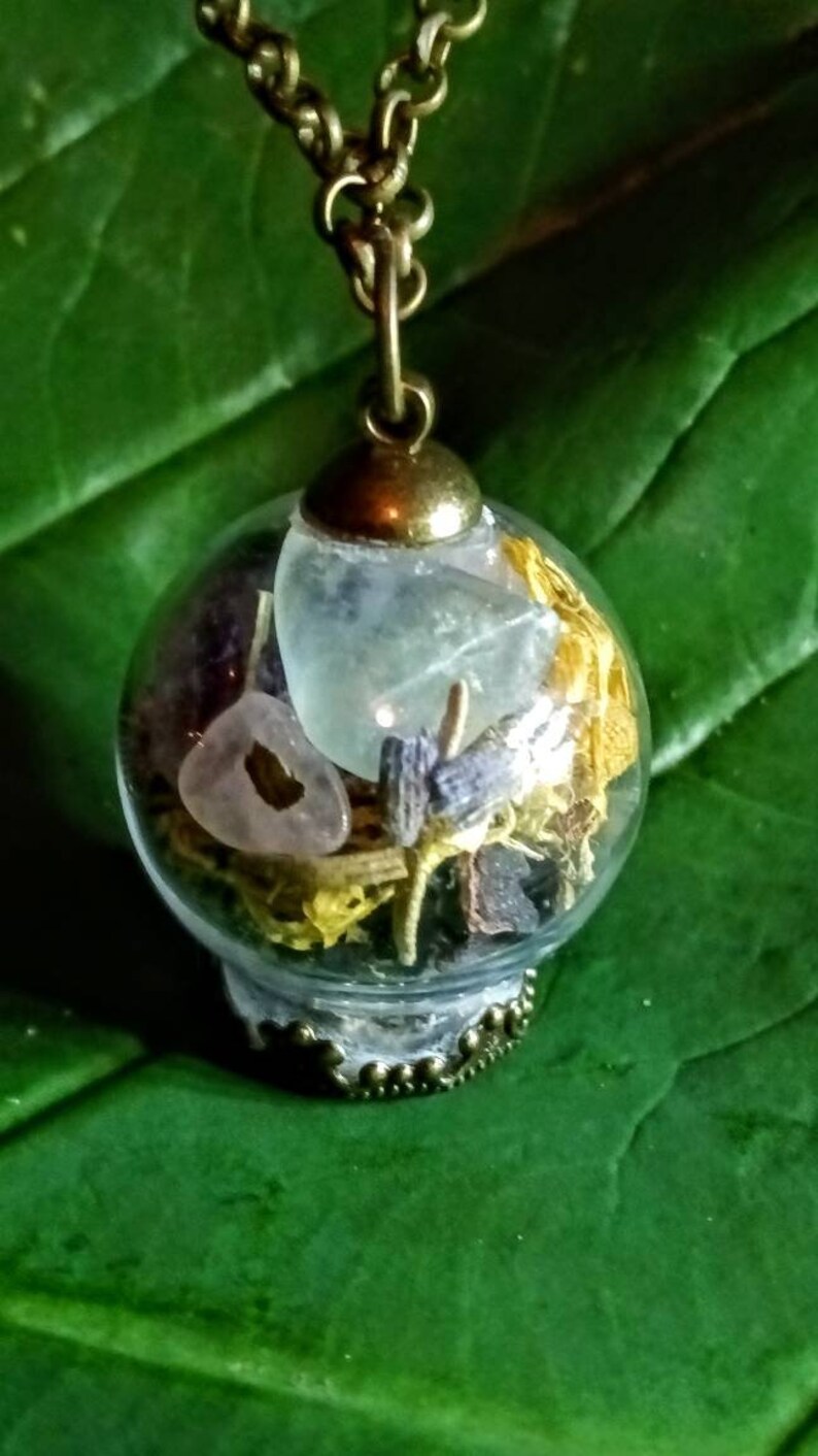 May include: A gold chain necklace with a glass orb pendant filled with dried flowers, crystals, and herbs. The orb is clear and has a gold-toned metal rim.