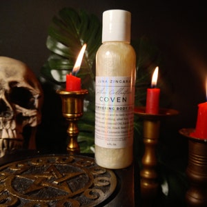 May include: A clear glass bottle with a gold shimmery liquid inside. The bottle is labeled "Luna Zingara The Collection Coven Shimmering Body Oil". The bottle is sitting on a black surface with a gold pentagram design. There are three lit candles in the background.