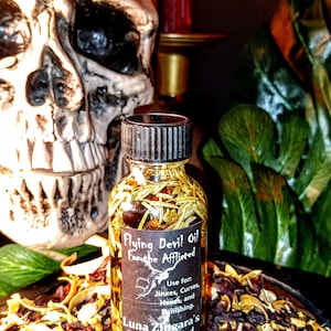 May include: A small glass bottle filled with a golden liquid and herbs. The bottle has a black label with the text "Flying Devil Oil For the Afflicted" and "Luna Zingara's Curiously Crafted Cauldron Creations". The bottle is sitting on a bed of dried herbs and flowers.
