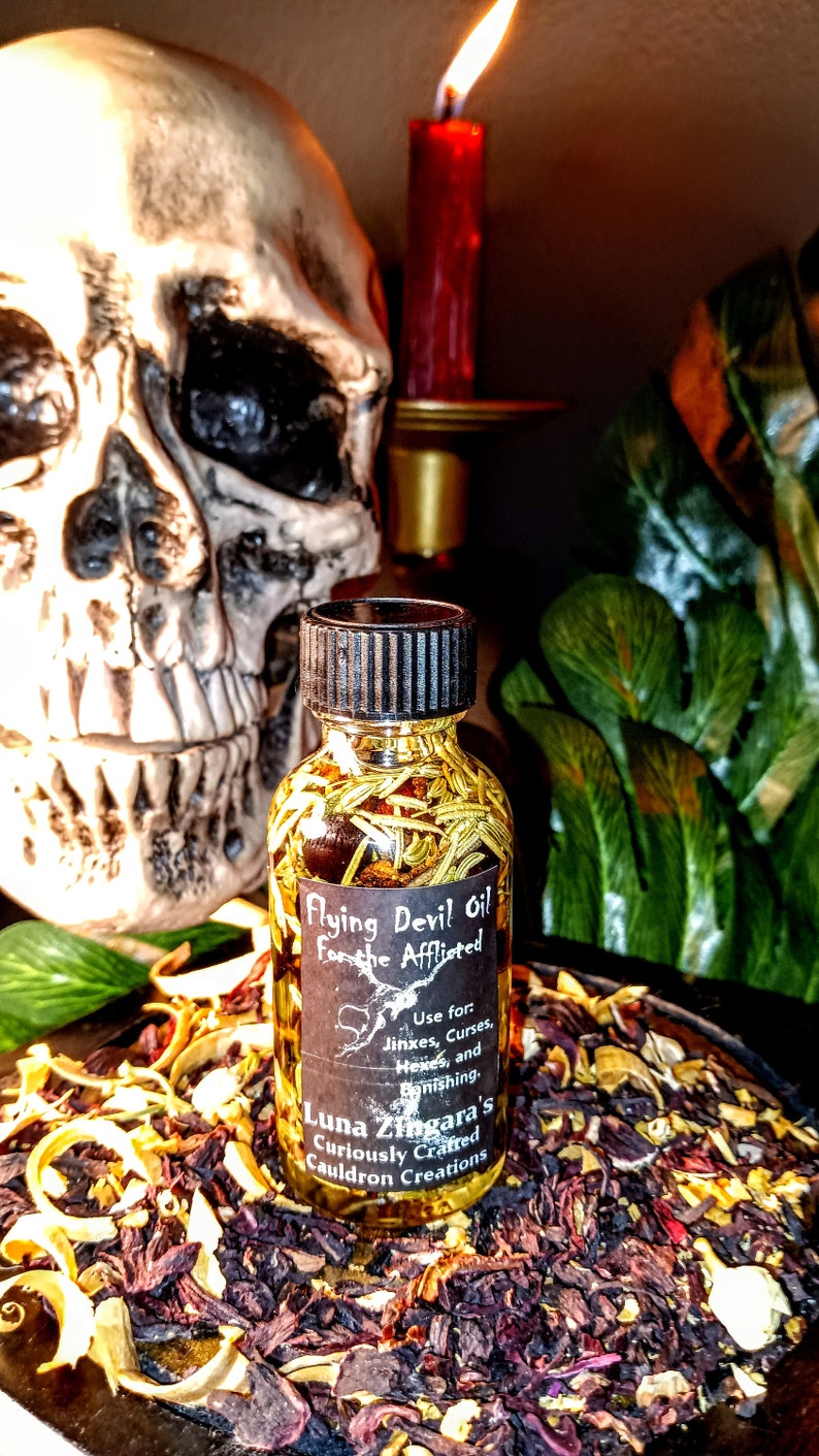 May include: A small glass bottle filled with a golden liquid and herbs. The bottle has a black label with the text "Flying Devil Oil For the Afflicted" and "Luna Zingara's Curiously Crafted Cauldron Creations". The bottle is sitting on a bed of dried herbs and flowers.